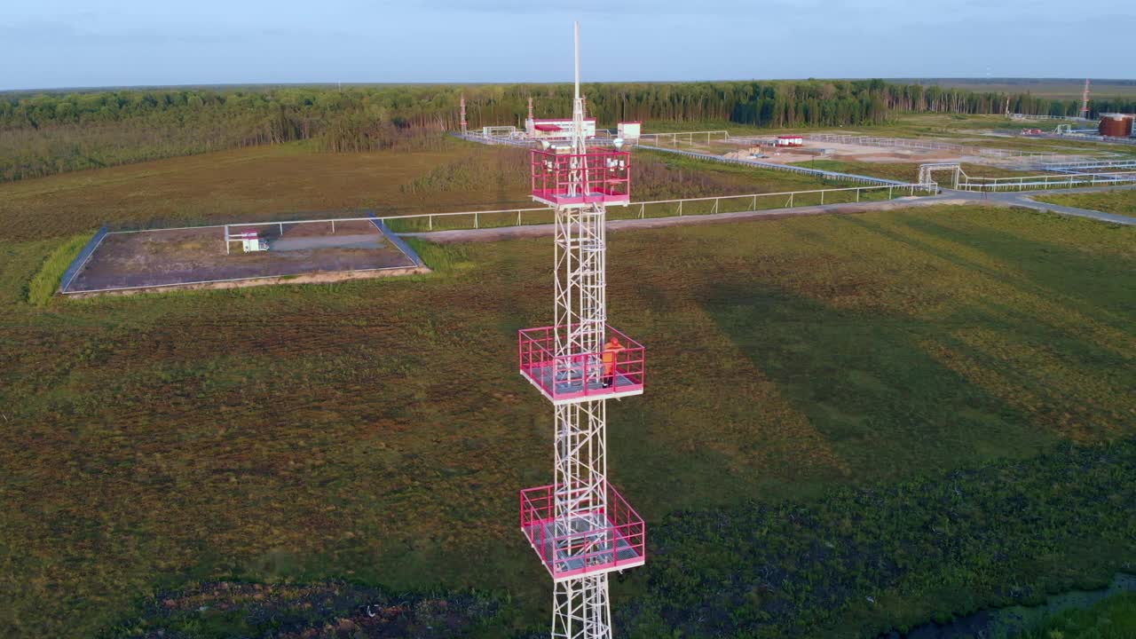 在落日的余晖中，一名正在工作的装配工站在高高的铁塔上，后面是西伯利亚沼泽中的油罐和油田。无人机飞到照明桅杆上向工人飞去视频下载