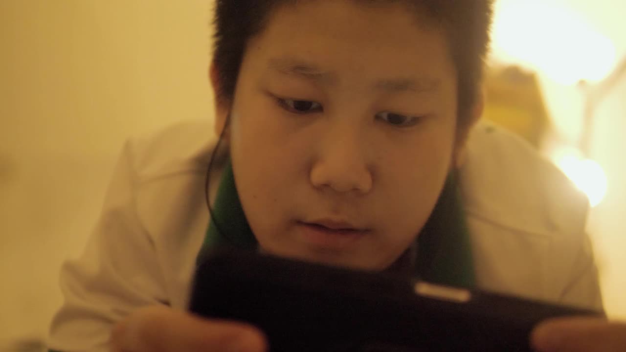 亚洲男孩晚上在床上用智能手机玩网络游戏，温暖的光线，生活理念。视频下载