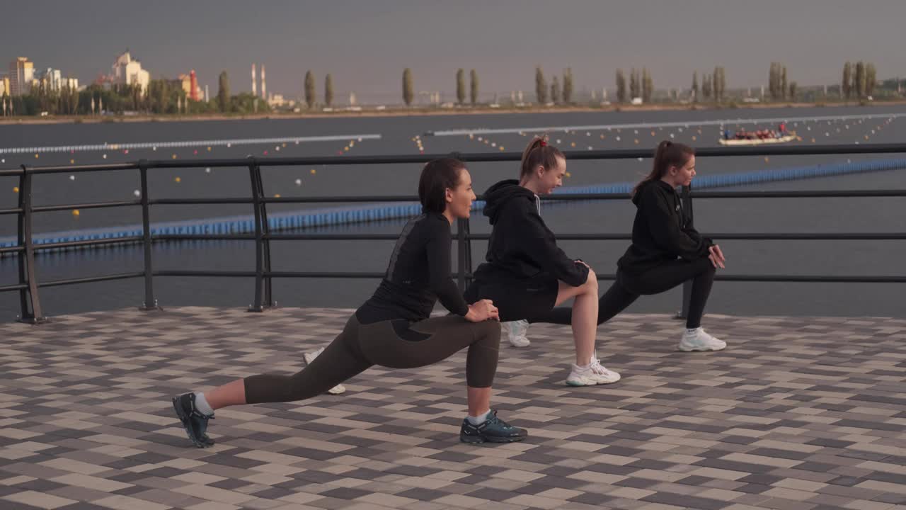 下午，三个运动女孩在河边的街道上做热身和伸展运动。女运动员视频下载