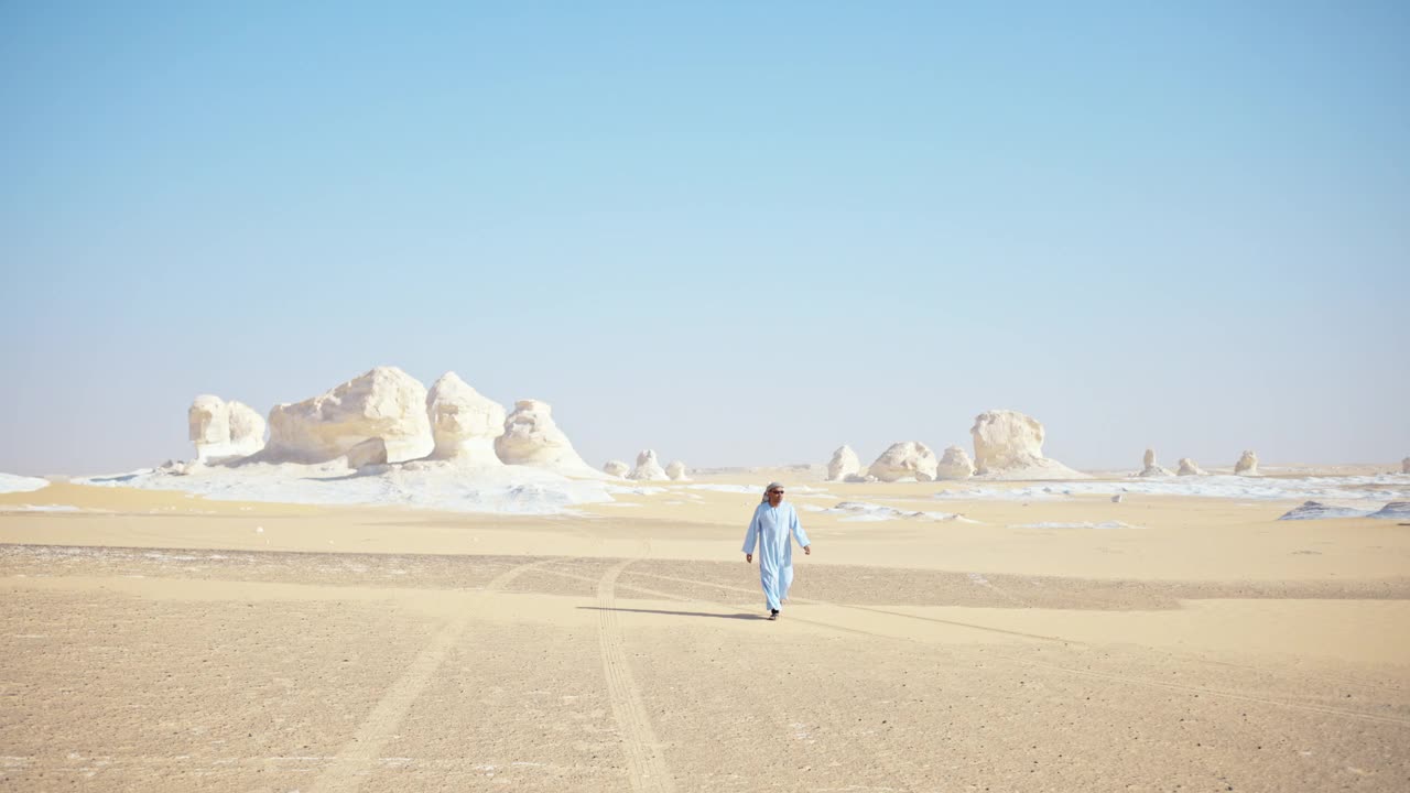 埃及游猎导游身穿浅蓝色传统长袍，头戴头巾，戴着黑色太阳镜，在超现实的白色沙漠国家公园散步。视频下载