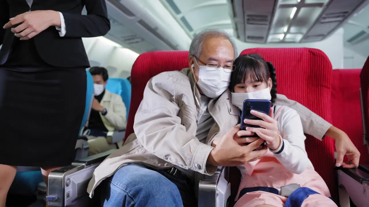 孙儿，亚洲少女戴着防护口罩用手机与亚洲爷爷和微笑，幸福地在飞机上出国新的旅程，其他乘客也用手机消磨无聊的时间。新常态旅行。视频下载