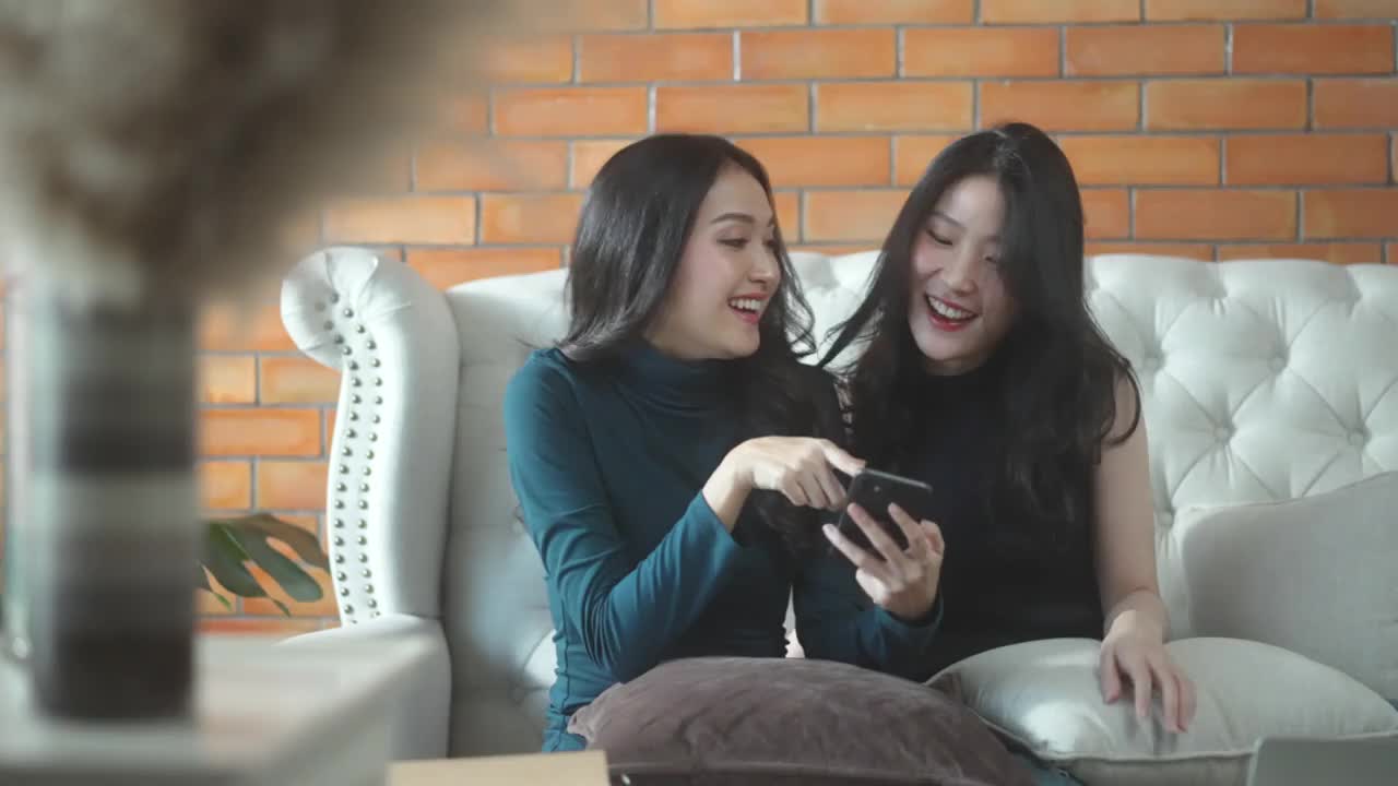 两个年轻的亚洲女性朋友一起分享快乐的时间，两个朋友放松与智能手机在沙发客厅视频素材