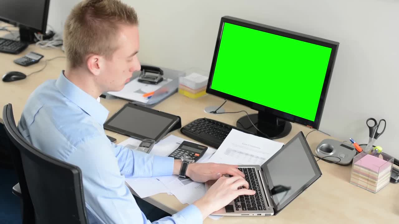 年轻英俊的男子在办公室用笔记本电脑工作-绿色屏幕-台式电脑和平板电脑视频素材
