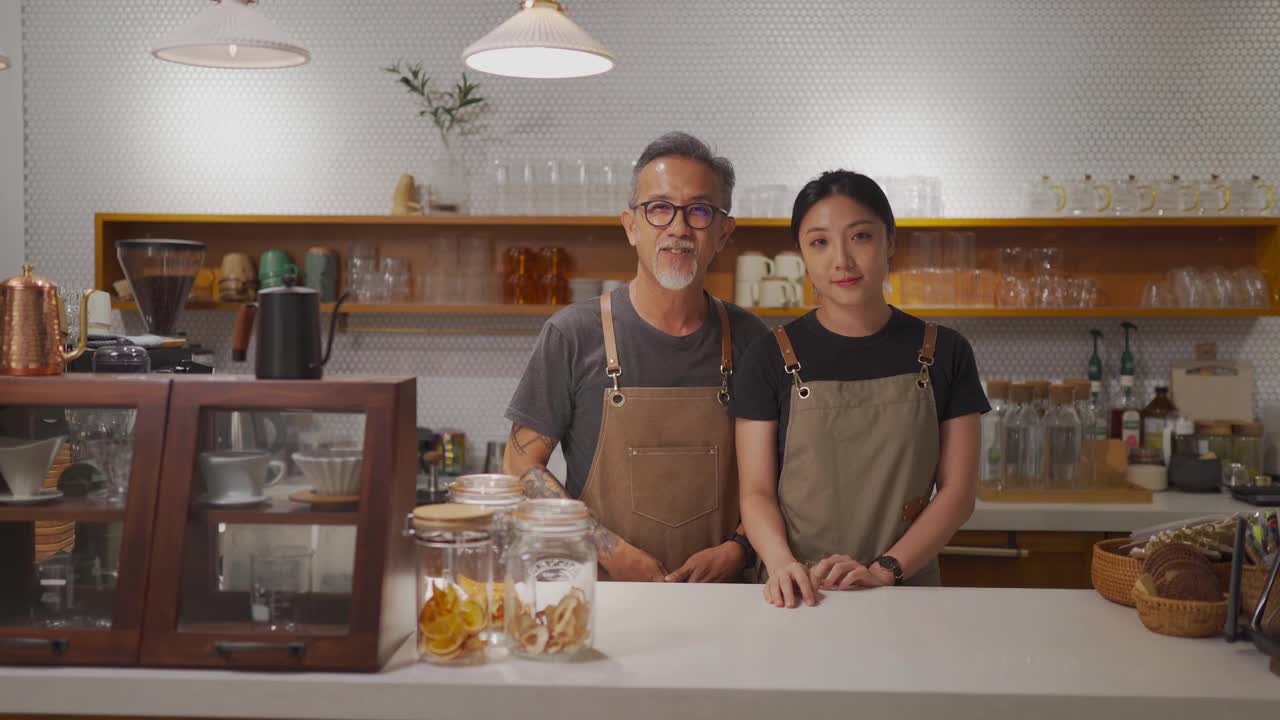 亚洲华人资深男性咖啡店老板和她的女儿看着镜头微笑在咖啡店柜台视频下载