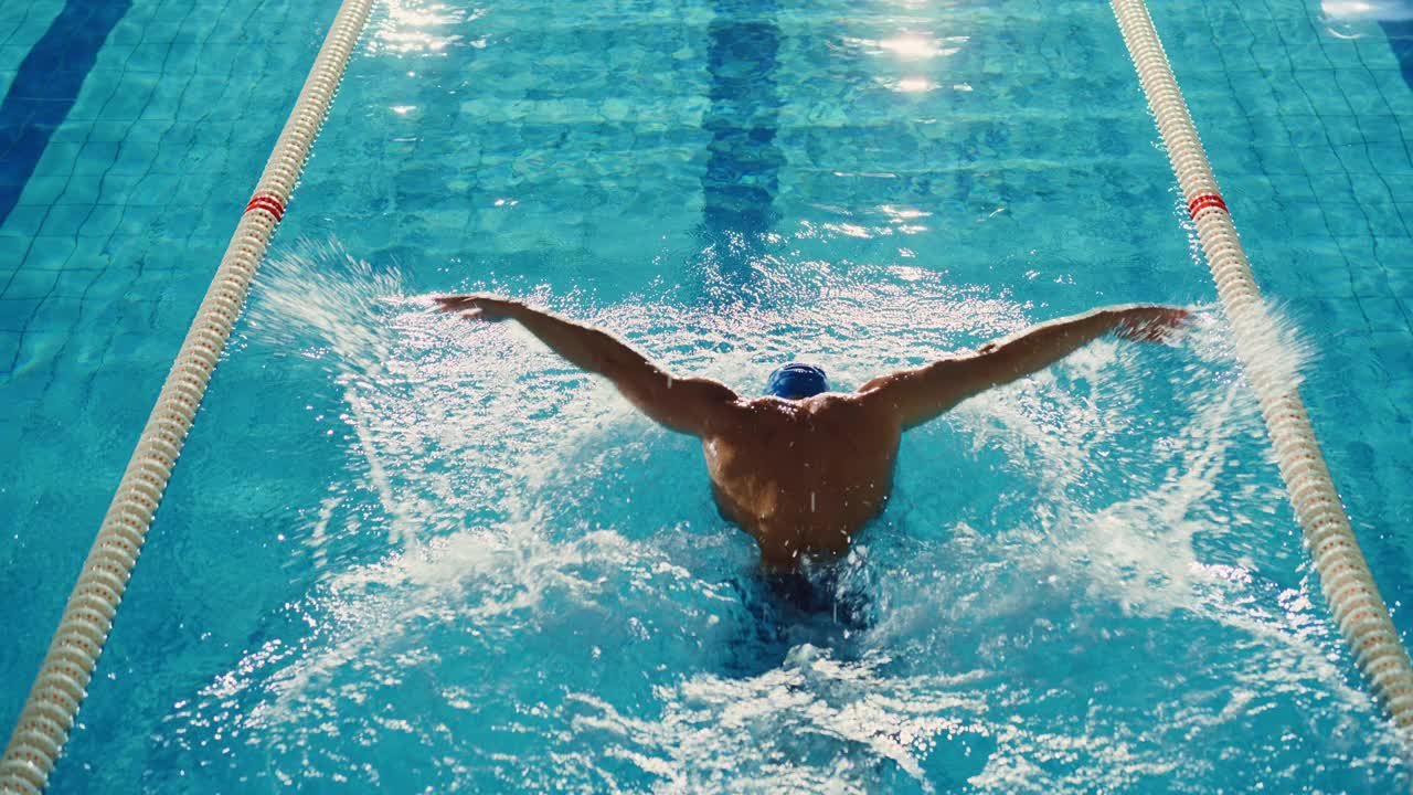 成功的男子游泳比赛，在游泳池游泳。决心用蝶式赢得冠军的职业运动员。丰富多彩的艺术电影风格。后视图空中跟踪镜头视频下载