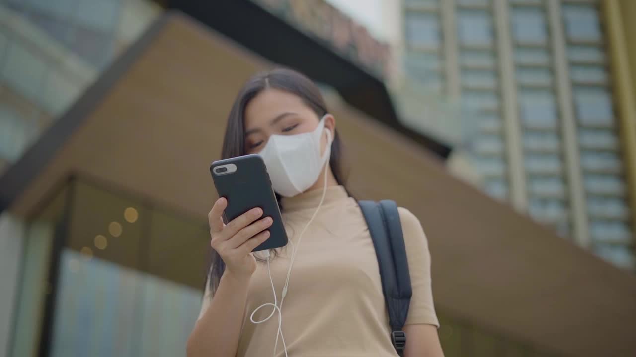 快乐的亚洲妇女戴着保护面罩和耳机使用智能手机听音乐走在城市街道上。视频素材
