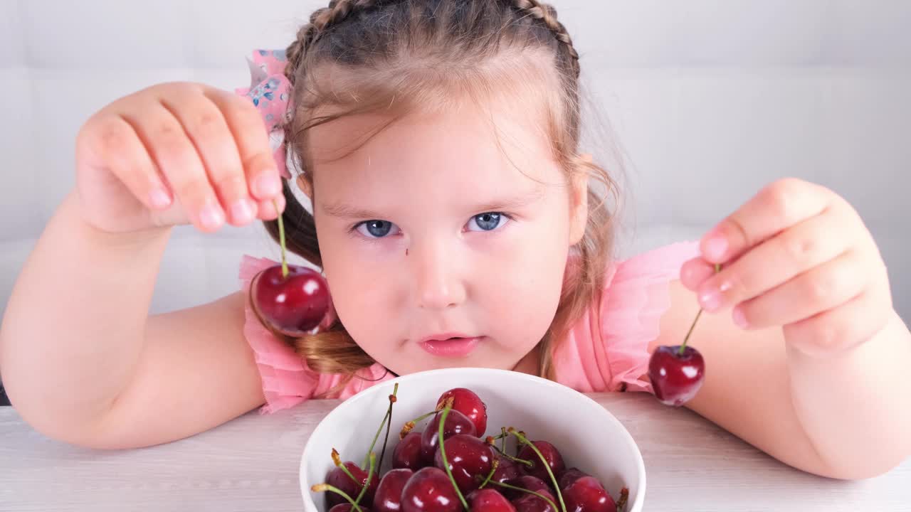 可爱的小女孩坐在一张轻木桌前，桌上放着一盘樱桃，手里拿着红浆果。健康饮食视频下载