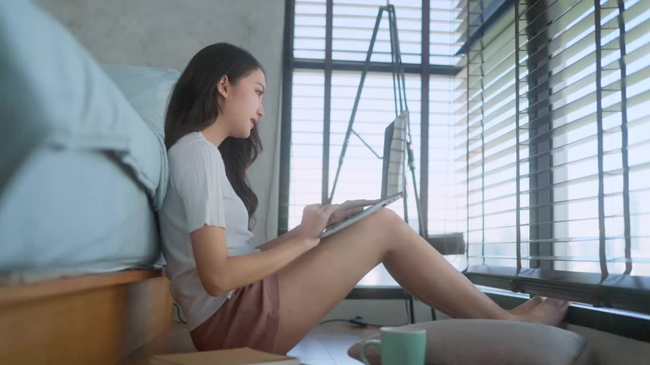 休闲亚洲女性穿着简单的布料工作与笔记本电脑旁边的大窗户在卧室的木地板上工作在家里的想法概念视频素材
