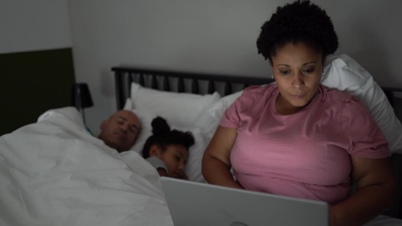 丈夫和女儿睡觉时，疲惫的女人在床上使用笔记本电脑视频下载
