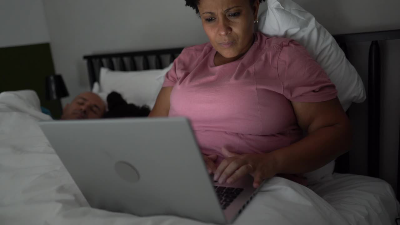 丈夫和女儿睡觉时，疲惫的女人在床上使用笔记本电脑视频下载