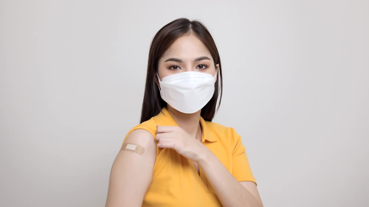 美丽的亚洲妇女穿着黄色衬衫戴着口罩，正在接种疫苗保护冠状病毒。快乐的女性在接种疫苗后用绷带指着手臂并竖起大拇指。视频素材
