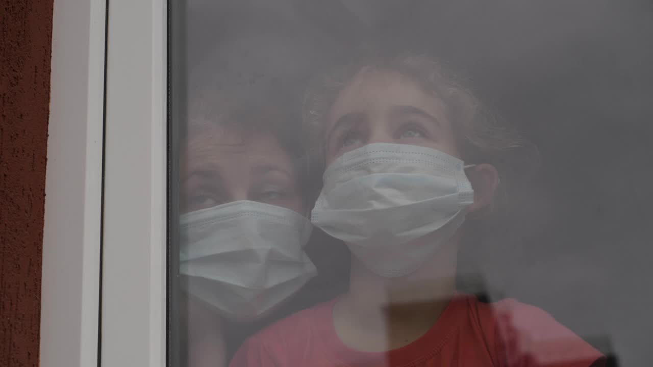 一名带着女儿的妇女，戴着防护口罩，悲伤地透过窗户看着，担心Covid-19被封锁。戴着防护面具的家人从窗户往外看。隔离冠状病毒社交距离视频素材