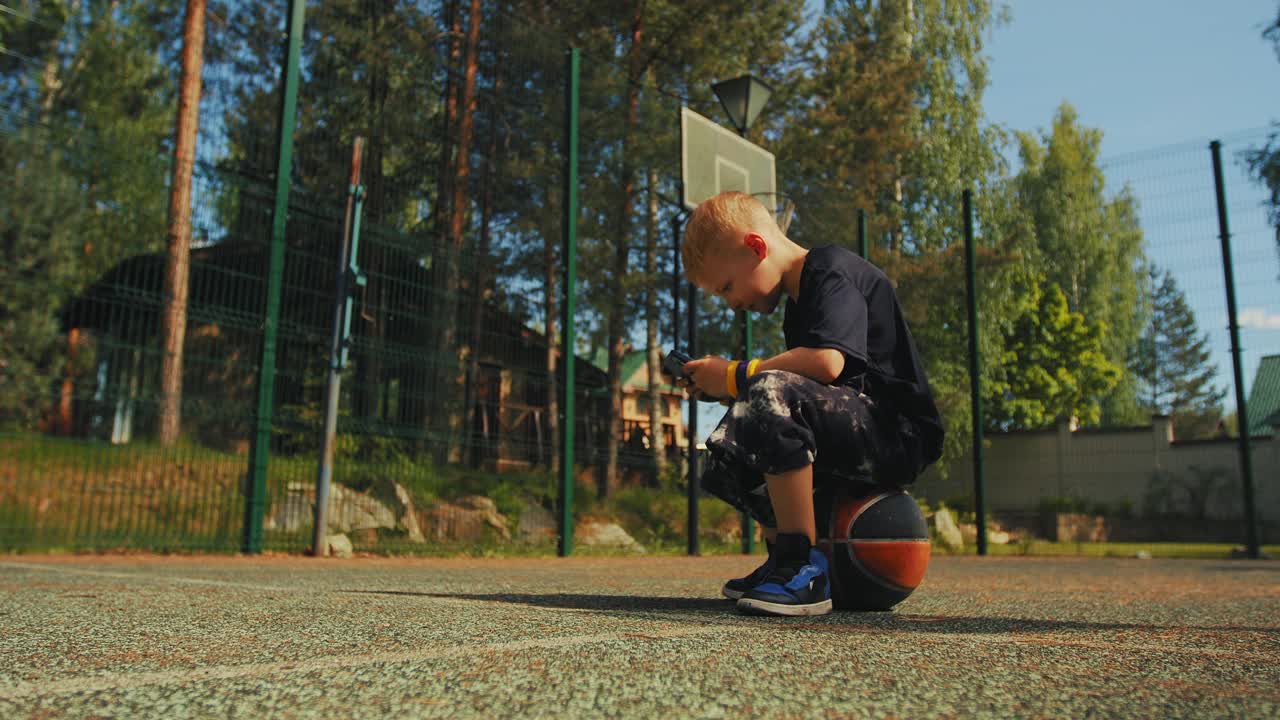 篮球运动员男孩坐在球上使用手机应用程序，看视频。一个男生在篮球场上玩网游视频下载