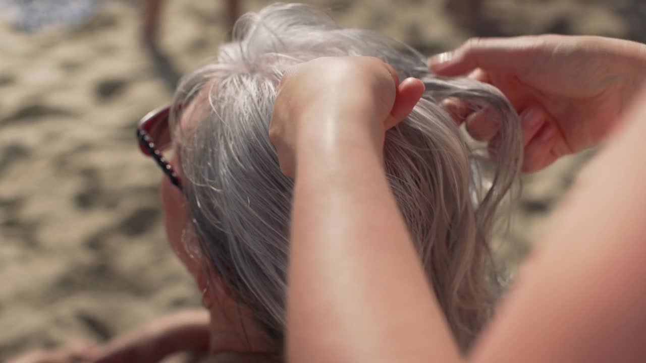 年轻女子编灰色头发，她年老的母亲休息在海滩上，近距离上的头和她的手，模糊的沙子背景视频下载