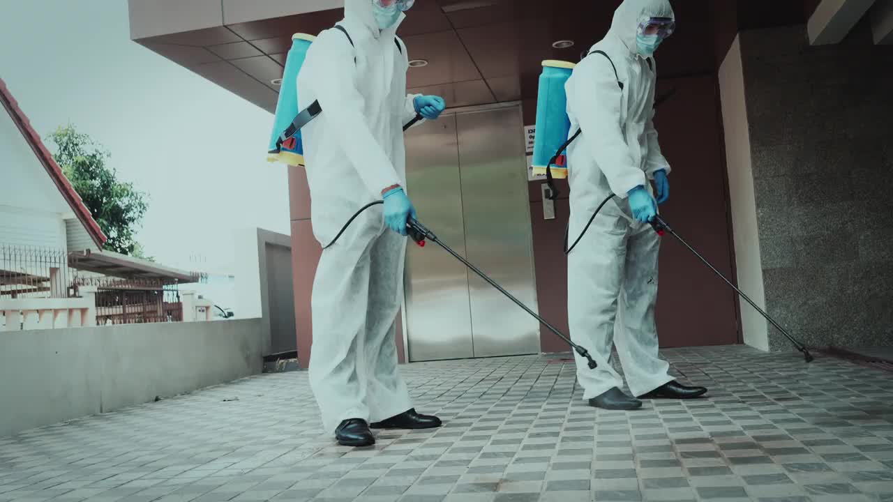 2名专家男性，身着防护服，戴口罩，在办公室和病原体滋生地喷洒消毒，4K分辨率，COVID-19效应，视频素材