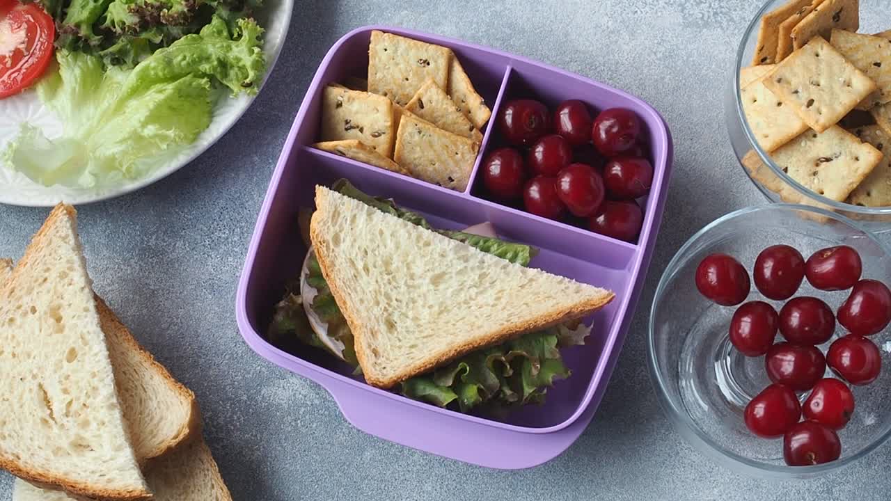女人把三明治放进午餐盒里。上学或上班的午餐，一个健康的零食三明治，樱桃和饼干。视频素材