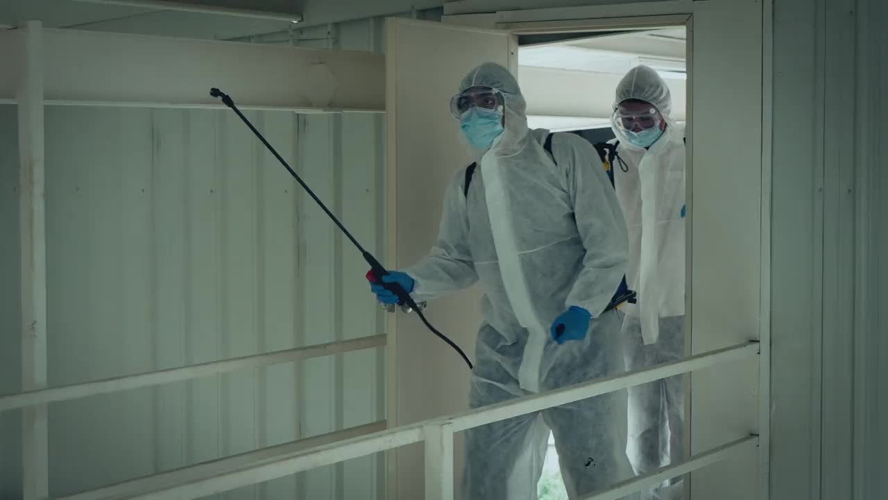 2名专家男性，身着防护服，戴口罩，在办公室和病原体滋生地喷洒消毒，4K分辨率，COVID-19效应，视频素材
