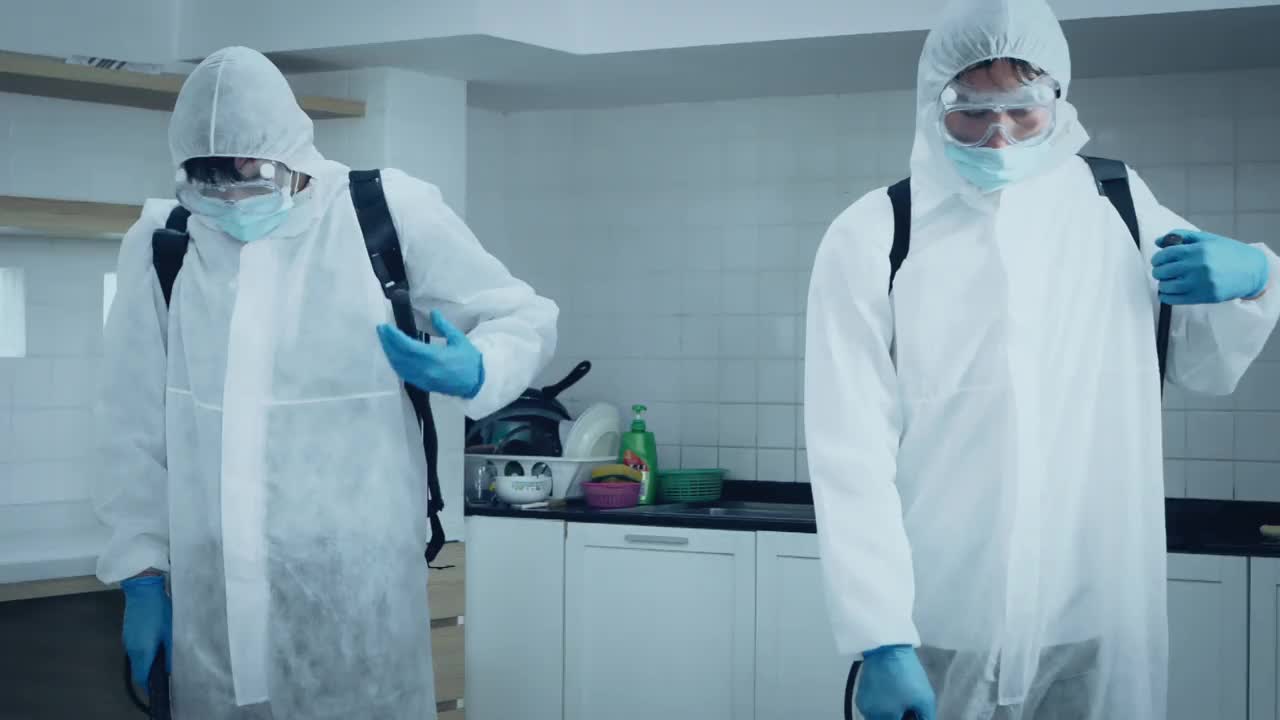 2名专家男性，身着防护服，戴口罩，在办公室和病原体滋生地喷洒消毒，4K分辨率，COVID-19效应，慢动作。视频素材