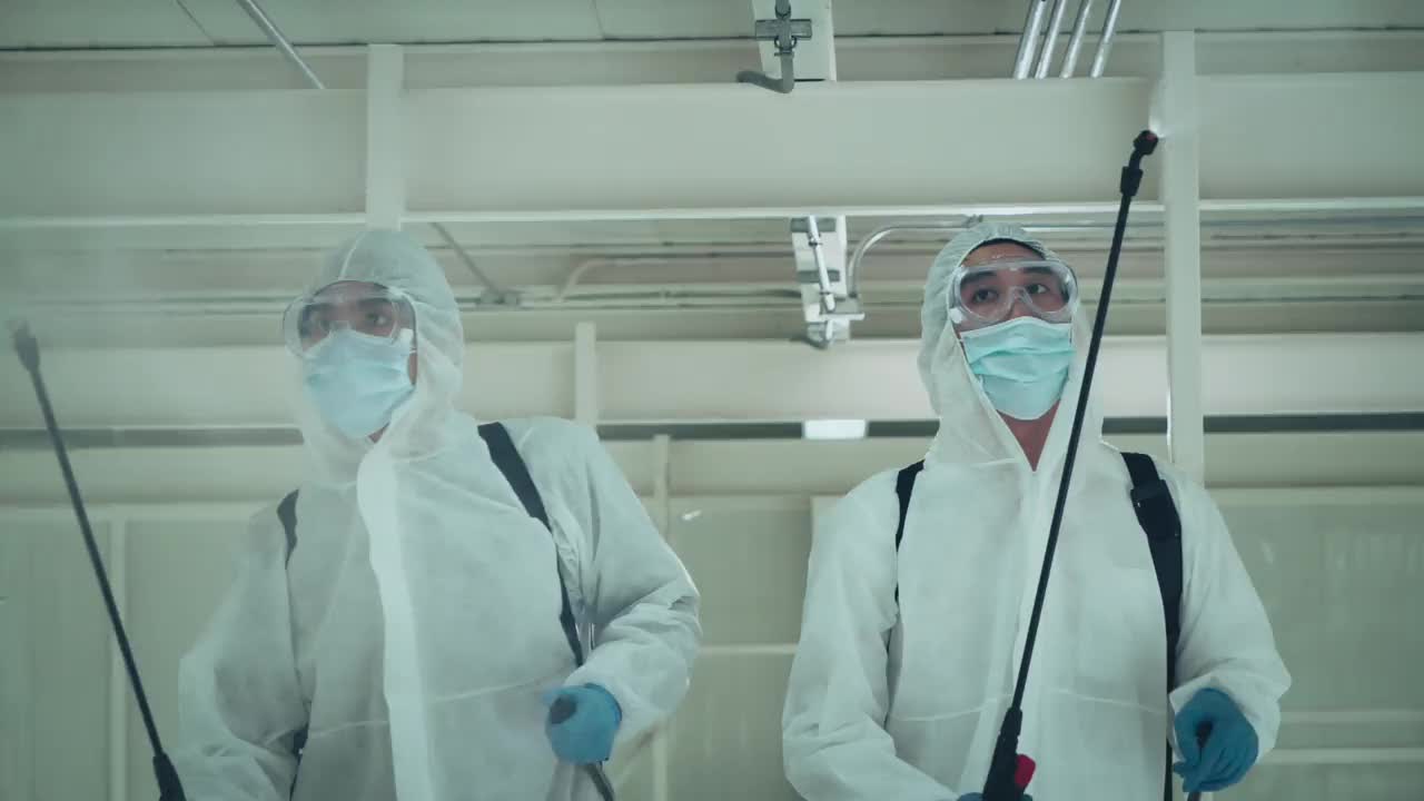 2名专家男性，身着防护服，戴口罩，在办公室和病原体滋生地喷洒消毒，4K分辨率，COVID-19效应，慢动作。视频素材