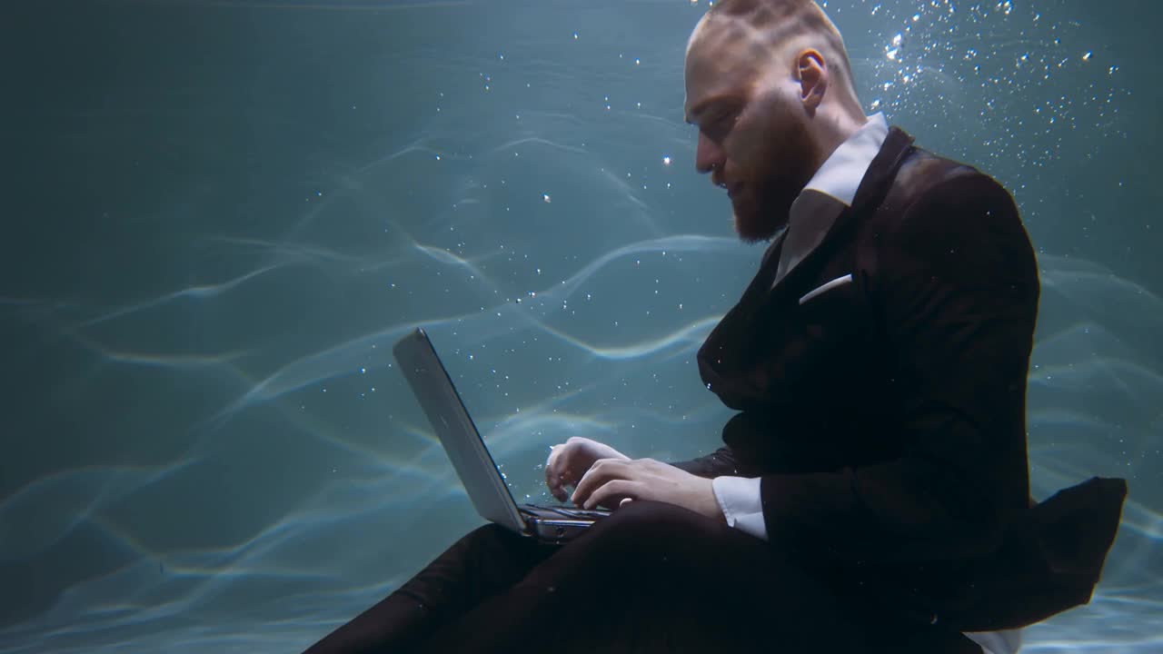 创造性的职业。电影在水下观看年轻快乐的商人西装思考，用笔记本电脑慢动作。视频下载