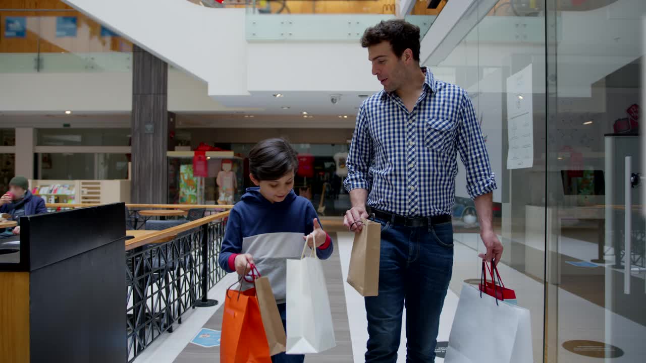 快乐甜蜜的男孩和他的爸爸在商场购物，每个人拿着袋子，同时愉快地交谈视频下载