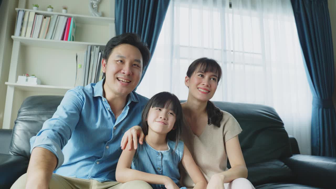 幸福的亚洲家庭呆在客厅里一起看电视电影。父亲和母亲花闲暇时间与小女孩女儿坐在沙发上，看动画片，在家里一起欢笑。视频素材