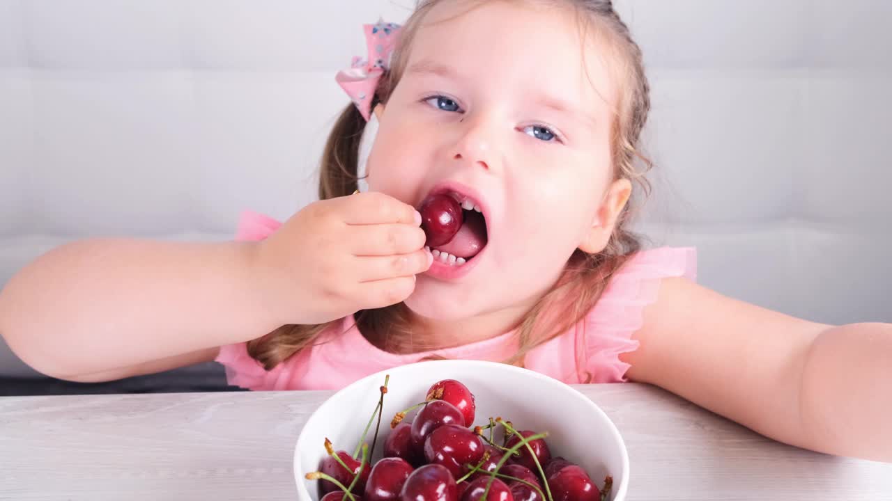 漂亮可爱的金发小女孩坐在一张轻木桌旁，桌上放着一盘樱桃，正在吃红浆果。健康饮食视频下载