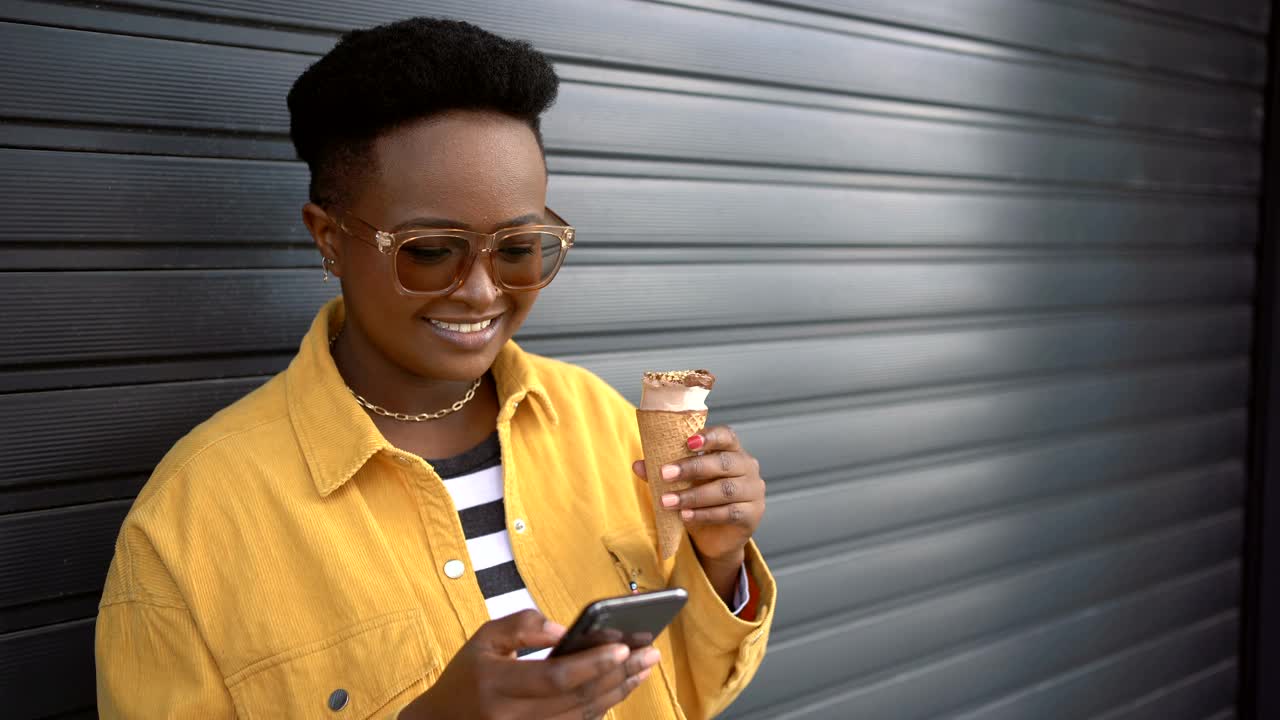 年轻，可爱的黑人妇女倚靠在深灰色车库门的背景，吃着冰淇淋视频下载