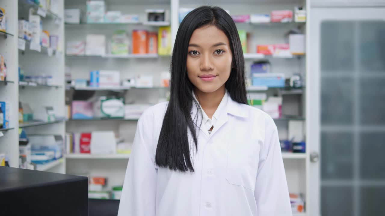 在医院药房工作的女药剂师肖像。快乐的年轻亚洲女医生穿着白色的医疗外套，看着相机。有药店药学背景的货架。视频下载