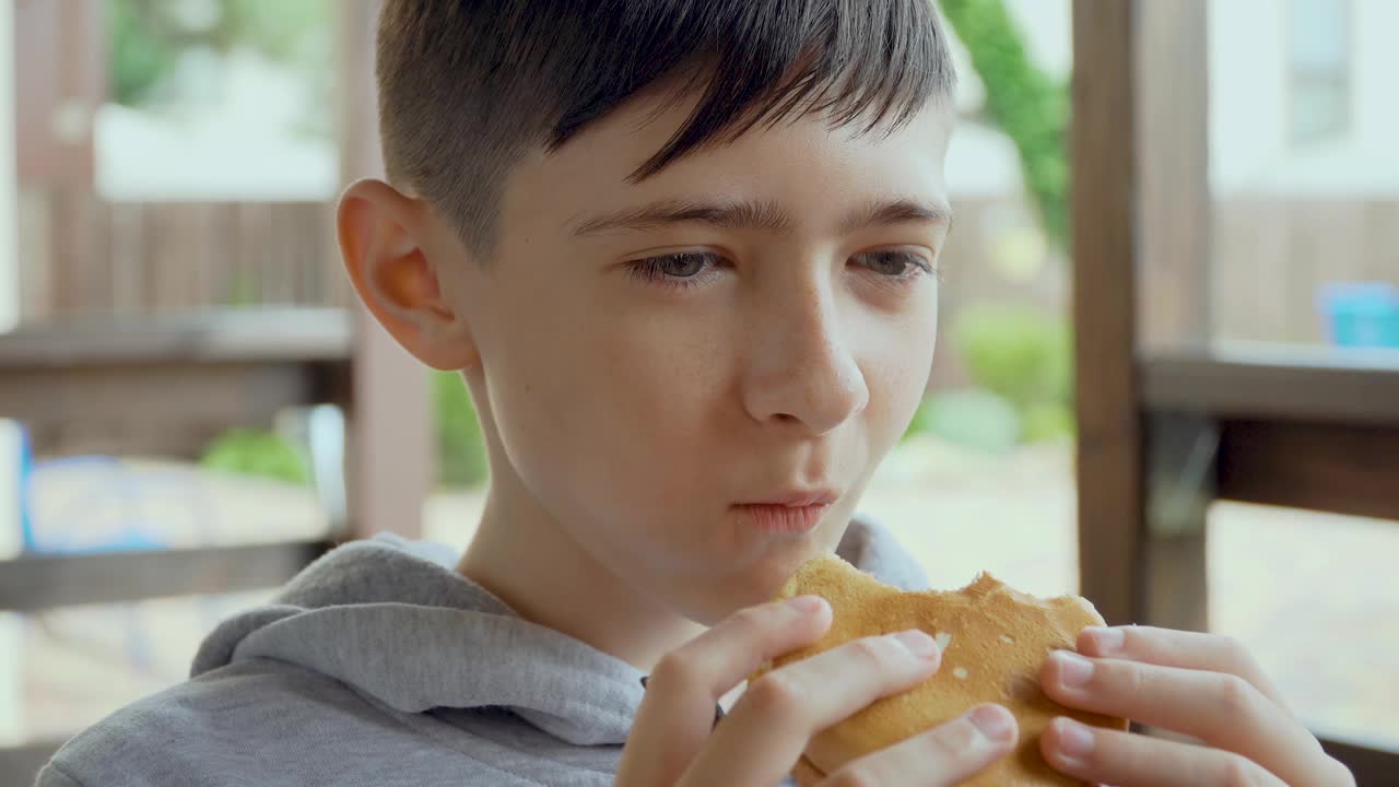 可爱的十几岁的男孩正坐在房子前面的凉亭里吃着快餐汉堡视频素材