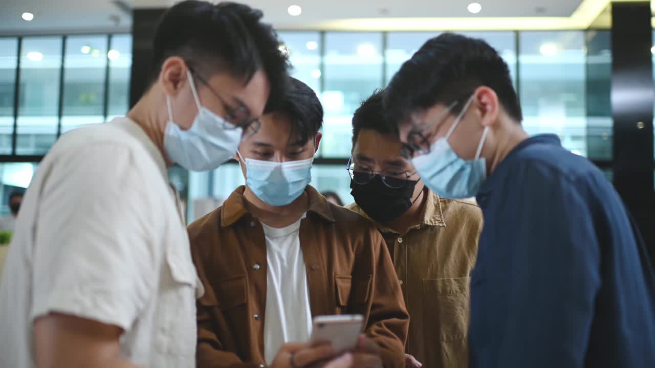 亚洲华人年轻人在电影院前戴着口罩看智能手机，用手机app在线订票视频下载