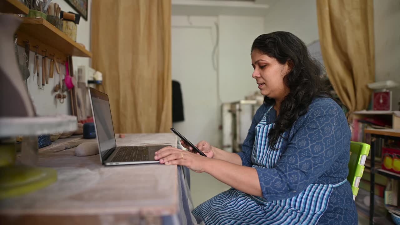 一名陶艺女艺术家在她的工作室里一边用笔记本电脑工作一边打电话视频素材