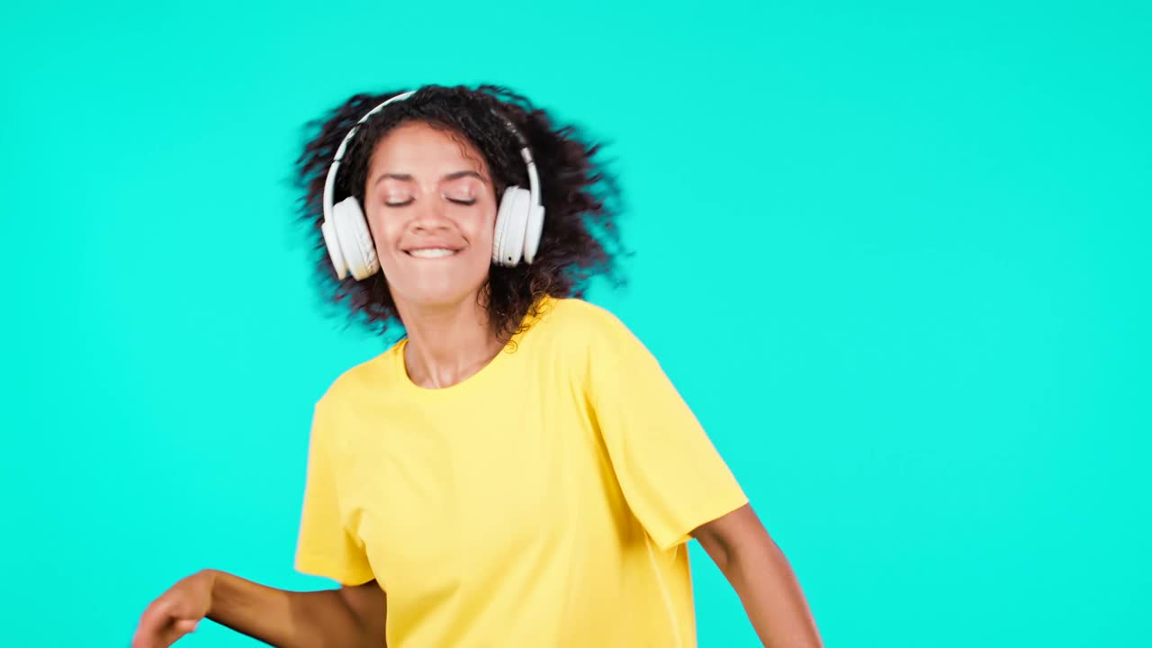 迷人的女人跳舞与无线耳机在蓝绿色的工作室背景。可爱的非洲裔美国女孩跳舞和微笑。音乐、广播、快乐、自由、青春理念。视频下载