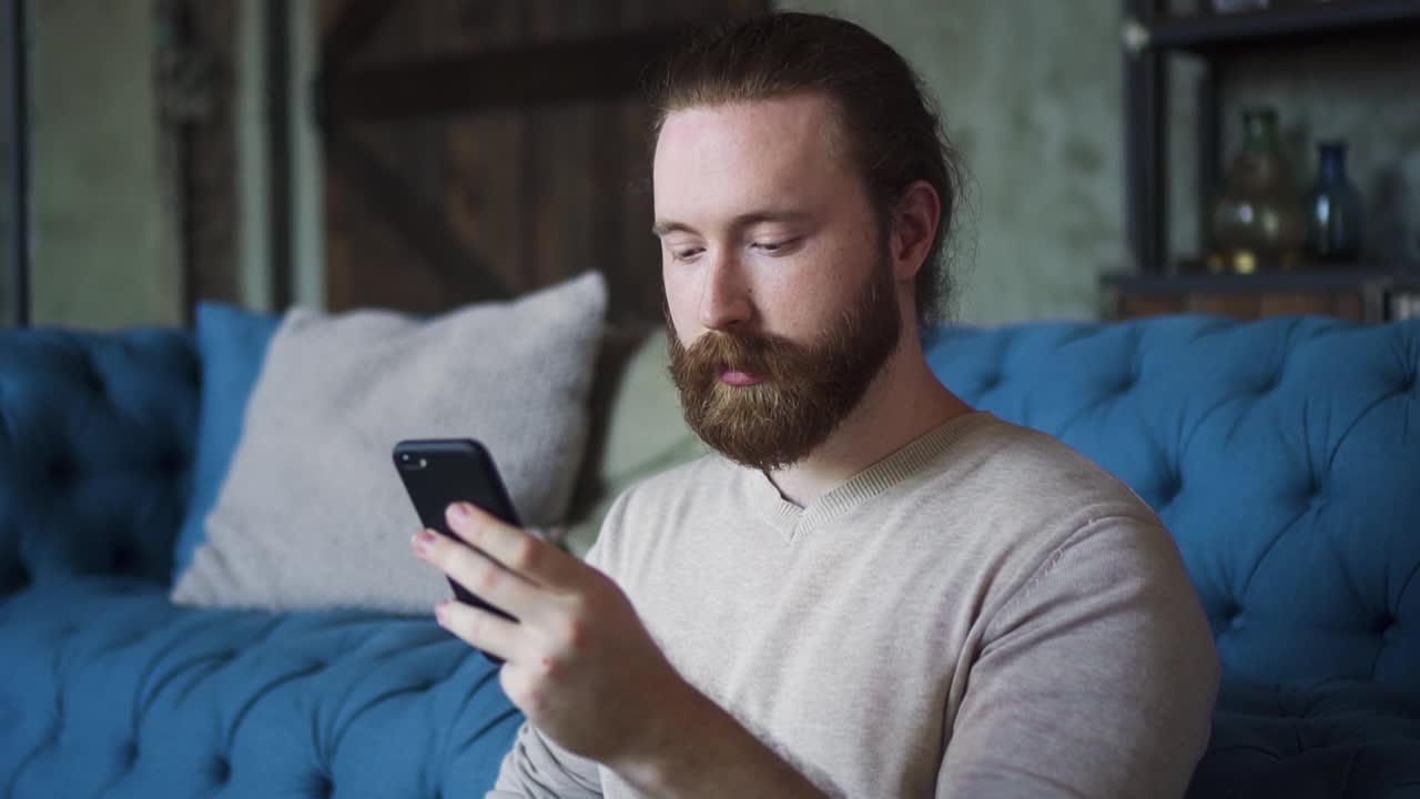 一个留着胡子的年轻人正在使用智能手机，在公寓房间里玩得很开心视频素材