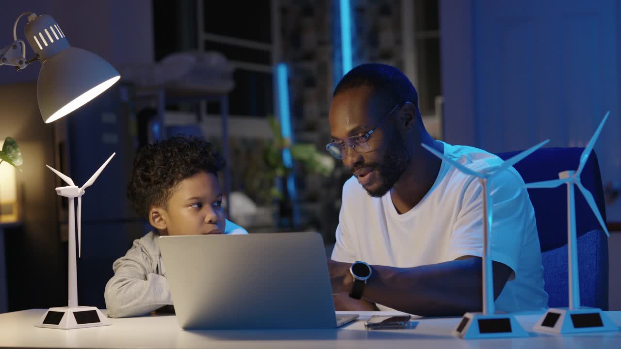非洲的父亲和9岁的儿子教生态学和讨论解释替代能源一起用笔记本电脑和模型涡轮机在晚上在家里。向孩子传授可持续发展知识视频下载