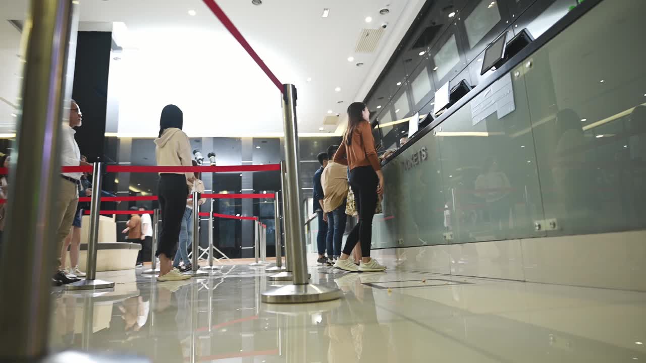 低角度观看亚洲华人观众在电影院售票柜台前排队购买电影票和爆米花饮料，在电影院放映前进行非接触式支付视频素材
