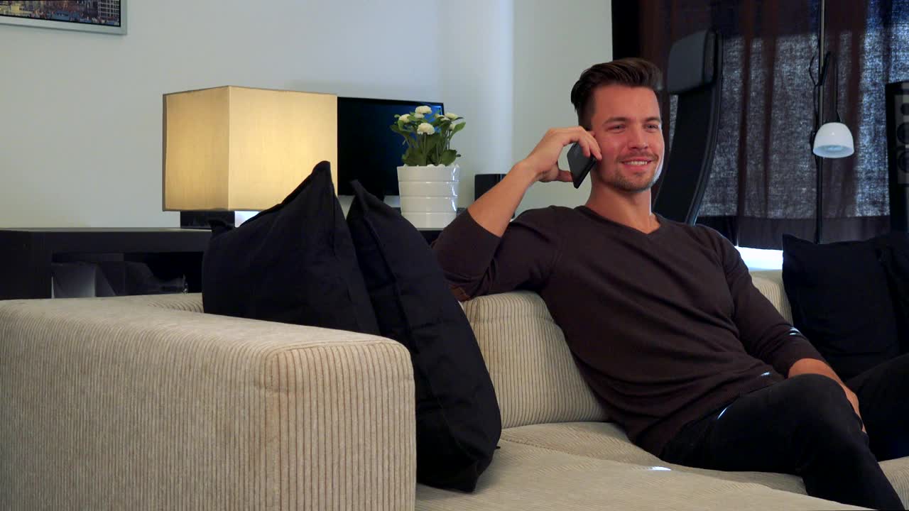 一个年轻英俊的男人坐在沙发上一边用智能手机聊天视频素材