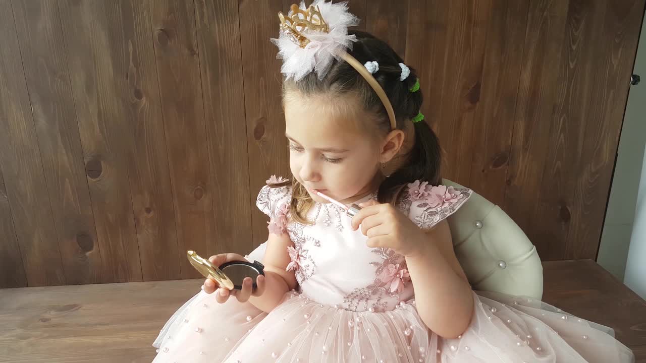 一个三岁的小女孩穿着华丽的粉红色裙子，头上戴着箍和皇冠，坐在儿童椅上，用口红涂着嘴唇，看着一面小镜子。视频下载