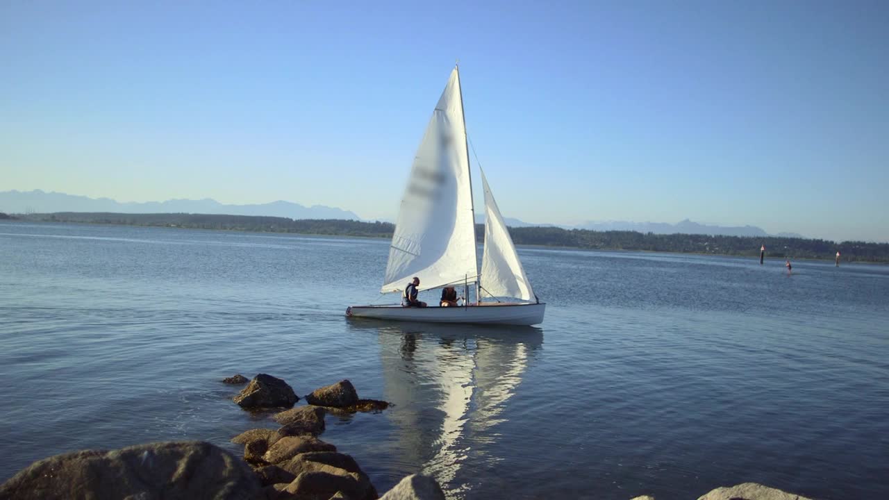 在明亮的蓝天和遥远的山脉中沿着湖边航行的小帆船-加拿大不列颠哥伦比亚省视频下载