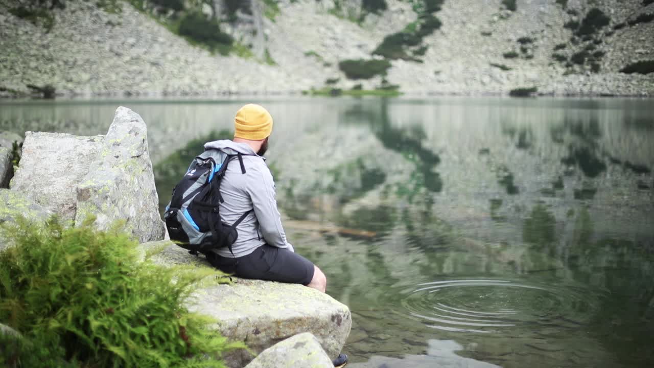 男游客坐在湖边的一块石头上，把石头扔进水里视频下载