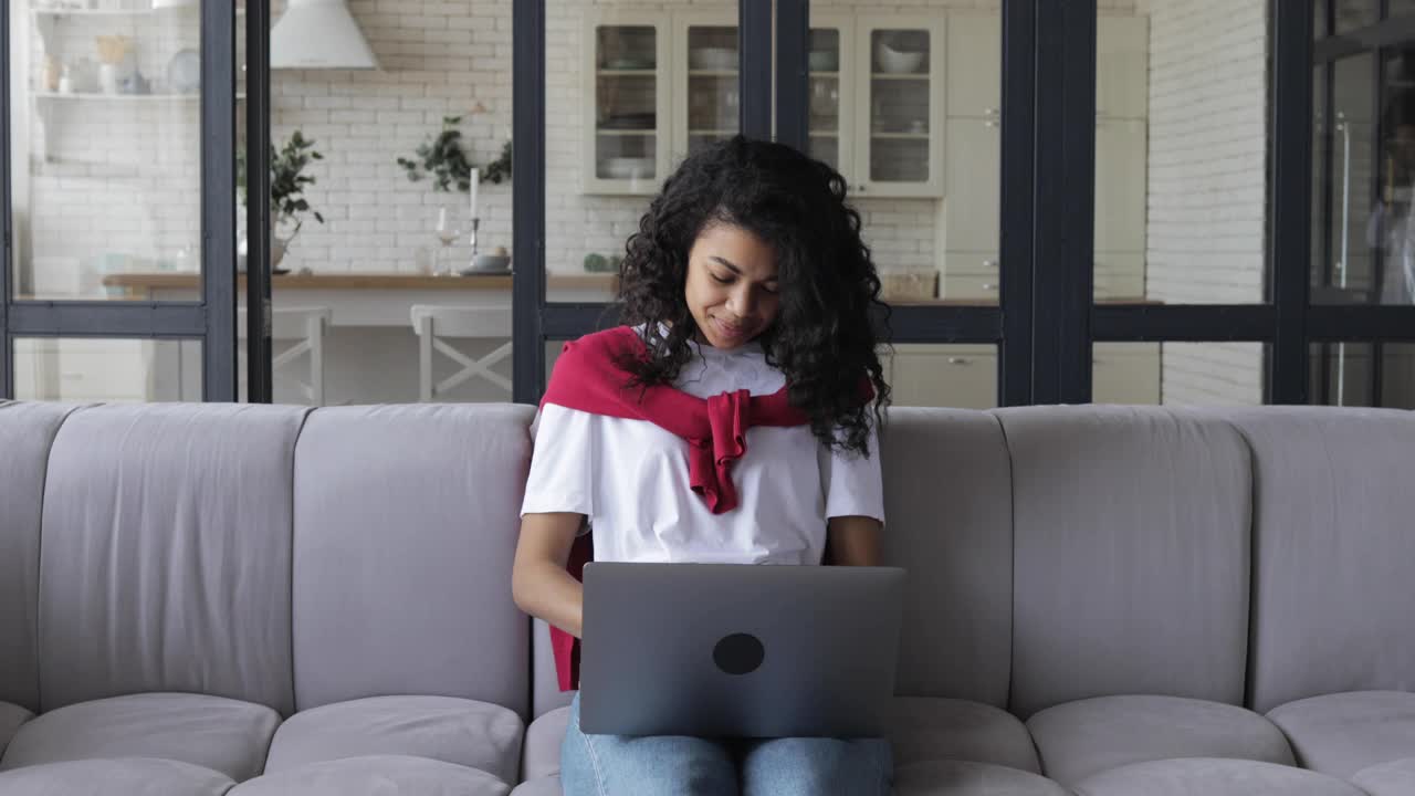一名年轻的非裔美国妇女坐在家里的沙发上，用笔记本电脑与一个正在上网的人交流并大喊大叫。咒骂并通过视频通话进行交流视频下载
