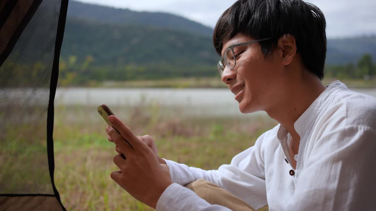 这是一名年轻男子在田园诗般的湖边露营时用智能手机发短信的侧面照片视频下载