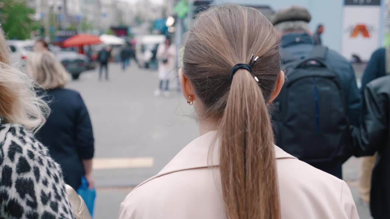 背影看到年轻时尚的女子穿着夹克走在市中心，在穿过马路穿过人群。模糊的人们从后面来到市中心，繁忙的城市生活视频下载
