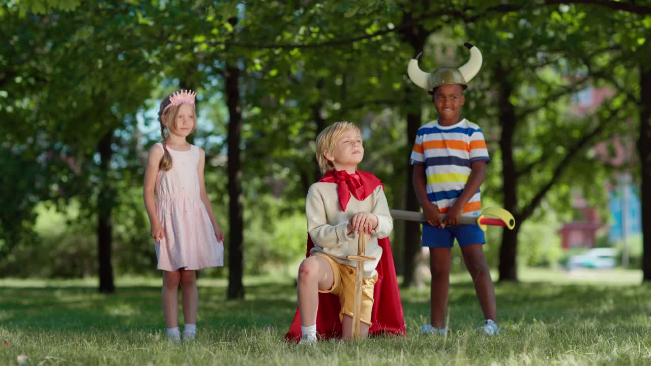 各种各样的孩子穿着戏服在公园里扮演角色。小男孩戴着红斗篷，跪在草地上，手里拿着剑。欣喜若狂的男孩举起手举着剑。孩子们为国王鼓掌视频下载