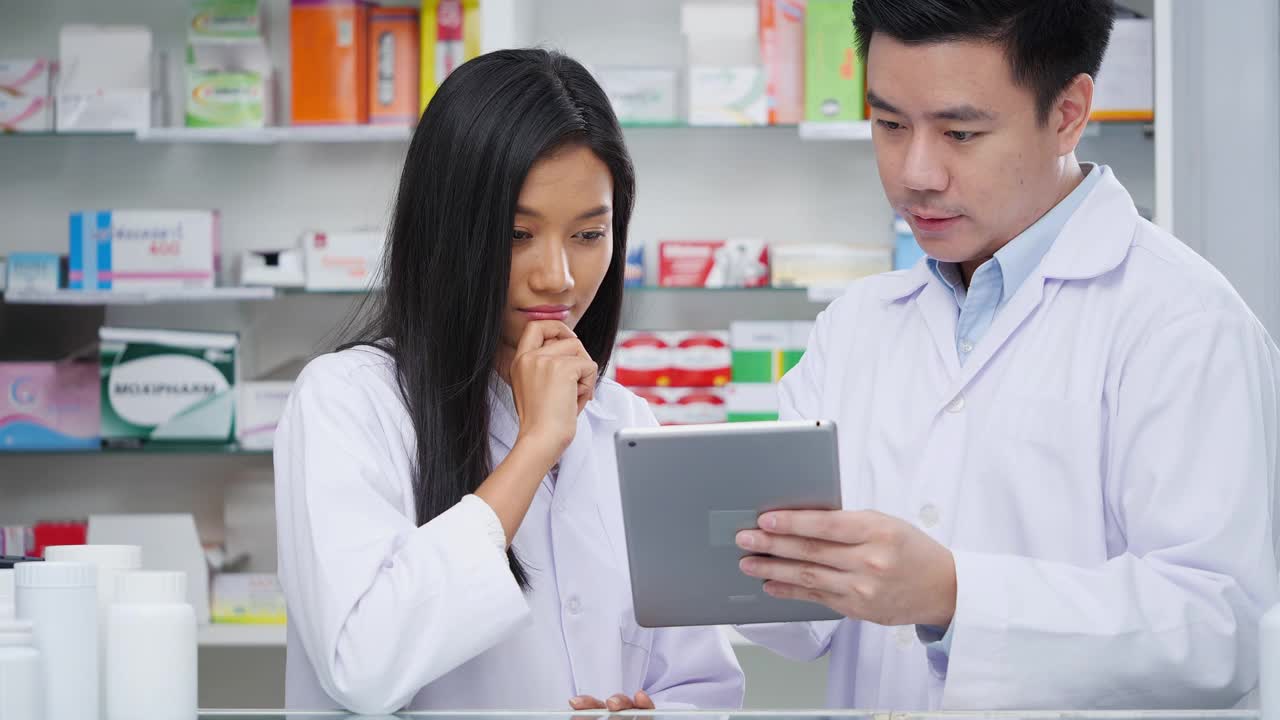 图为在医院药房检查药品的男女药剂师的照片。亚洲医生正在检查药片上的药物数据，穿着医疗外套。有药店药学背景的货架。视频素材