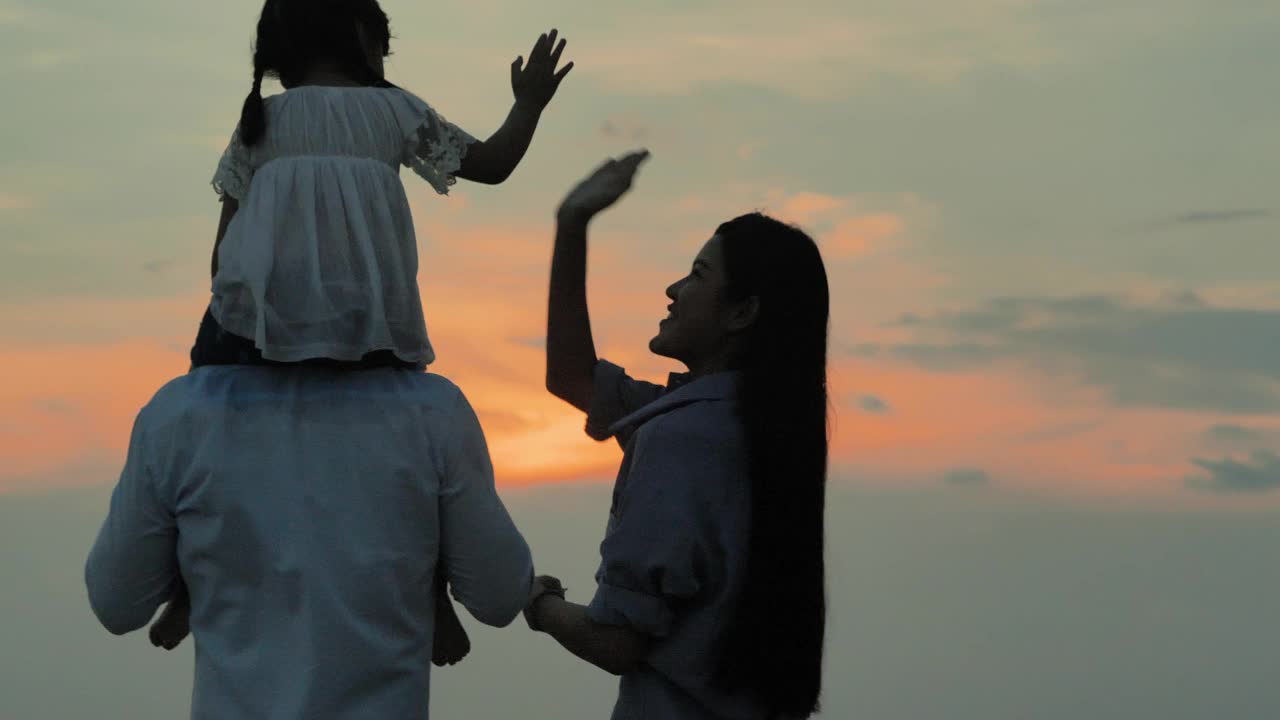 后视镜，一个4岁的亚洲女孩骑在父亲的脖子上，母亲牵着她的手，看着海滩上的日落。幸福的家庭假期。援助之手的概念。视频素材