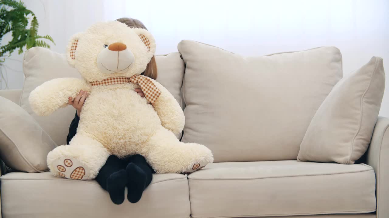 小女孩微笑着坐在沙发上，她的玩具泰迪熊在4k视频。视频素材
