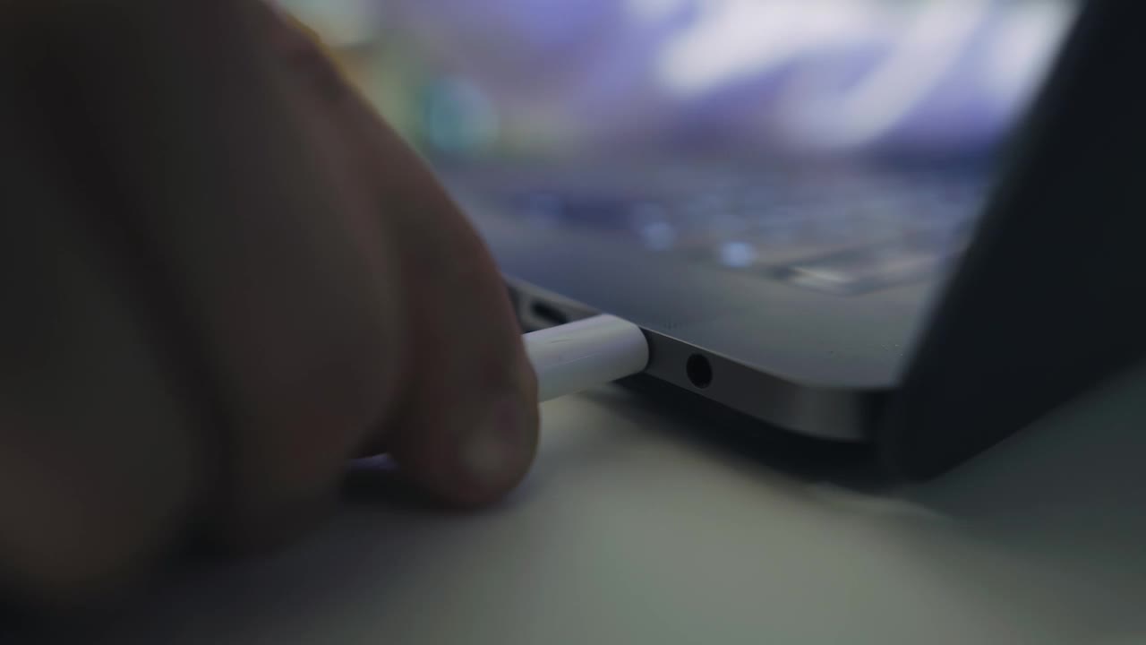 一名男子将一根白色电线插头插入桌上一台笔记本电脑的端口。手的特写镜头视频素材