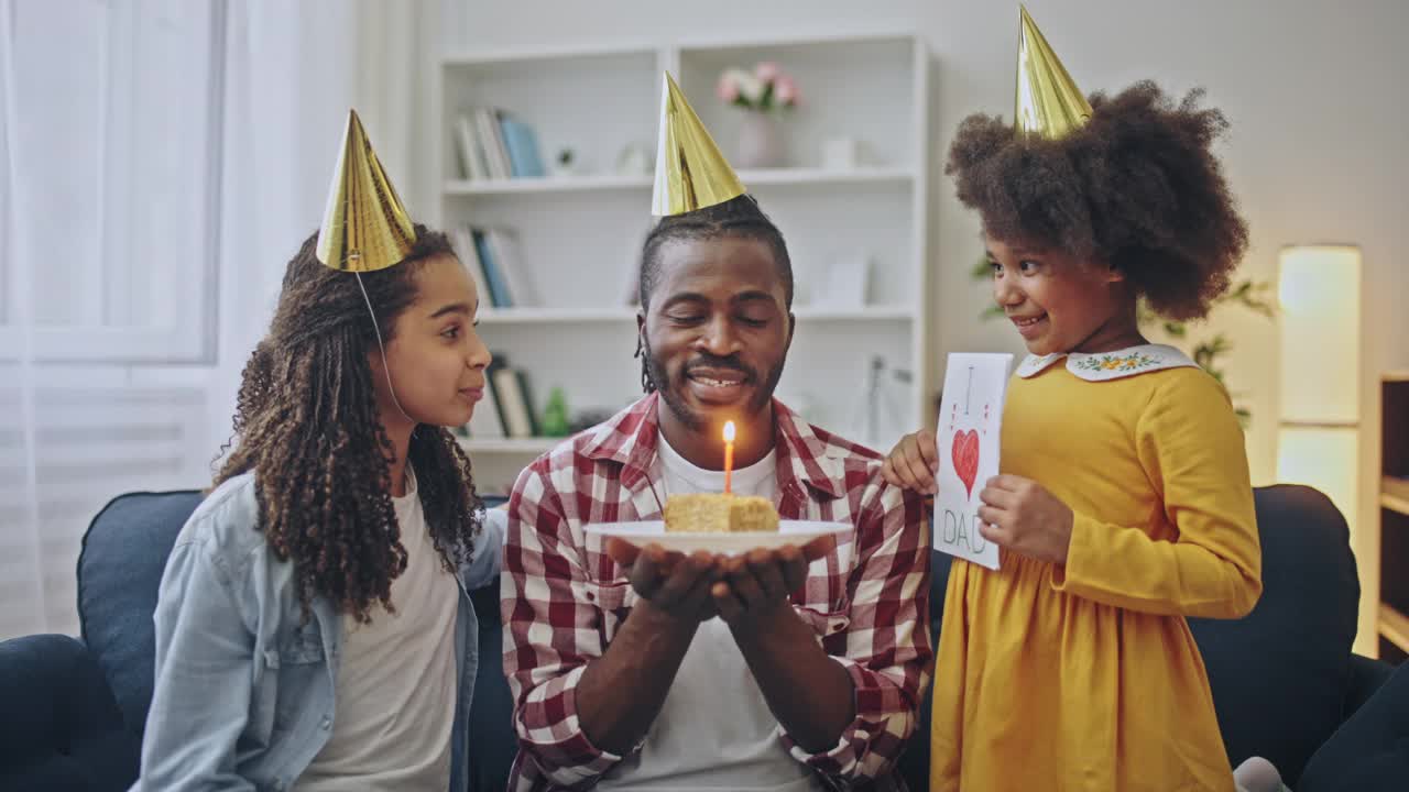 快乐的爸爸戴着派对帽吹着生日蛋糕蜡烛，和女儿们一起庆祝视频素材