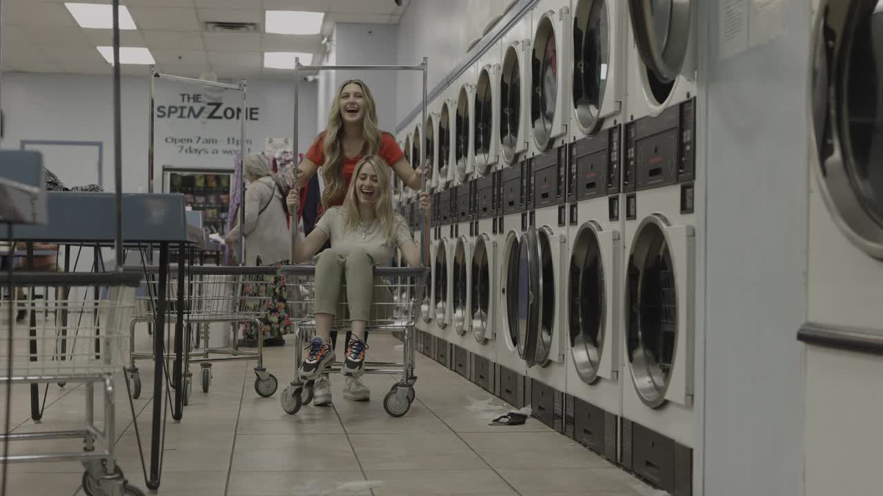 女孩在自助洗衣店推朋友坐在滚动的洗衣篮/普罗沃，犹他州，美国视频素材
